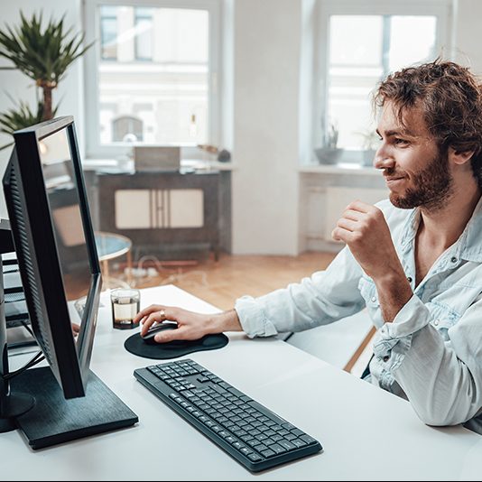 Joyful guy with curly hairs and beard sits at table working on pc